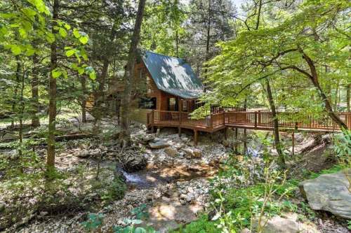 A cozy wooden cabin surrounded by lush greenery and a small stream, with a deck extending into the forest.