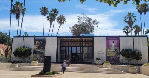 A modern building with large glass doors, surrounded by palm trees and decorative planters, under a clear blue sky.