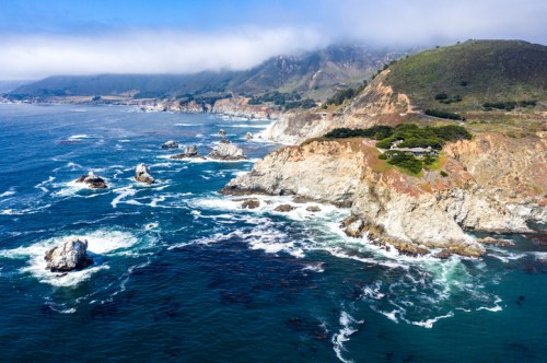 Aerial view of rugged coastline with rocky outcrops, waves crashing, and misty mountains in the background.