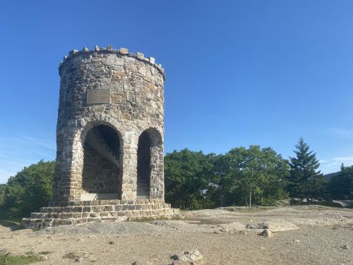 Stone tower with arched openings, surrounded by greenery and blue sky, on a rocky landscape.