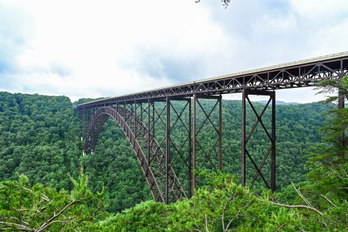 A large steel arch bridge spans a lush green valley under a cloudy sky.
