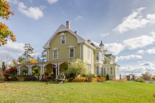 A large yellow Victorian-style house with a porch, surrounded by colorful autumn foliage and a clear blue sky.