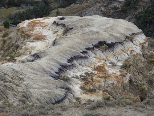 A textured rock formation with wavy patterns and patches of color, surrounded by sparse vegetation and hills.