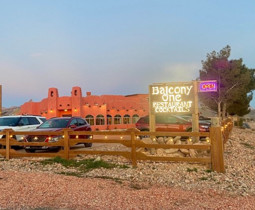 A colorful restaurant with a sign reading "Balcony One" and parked cars in front, set against a desert backdrop.