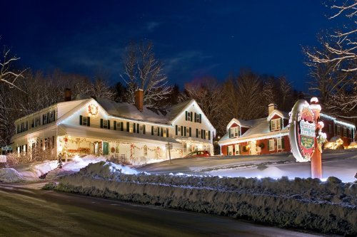 A charming, snow-covered house decorated with lights, surrounded by trees, under a twilight sky.