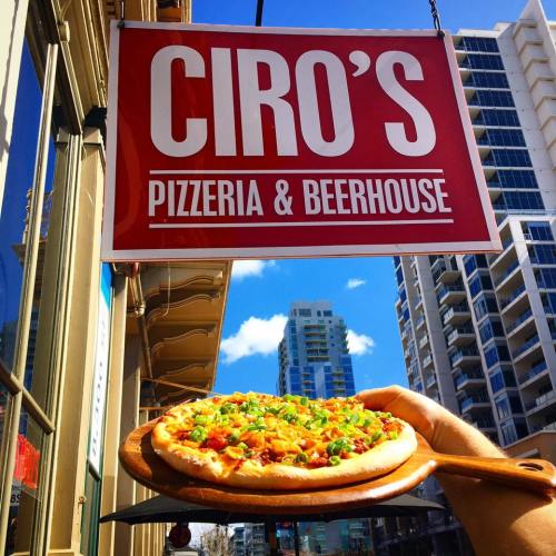 A hand holds a pizza topped with vegetables in front of Ciro's Pizzeria & Beerhouse sign against a city backdrop.