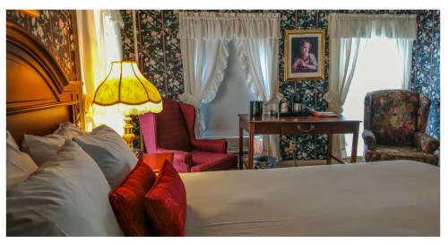 Cozy bedroom with a bed, floral wallpaper, a lamp, two armchairs, and a wooden desk with a portrait on the wall.