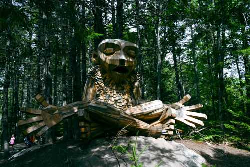 A large wooden sculpture of a seated figure with outstretched arms, surrounded by trees in a forest setting.