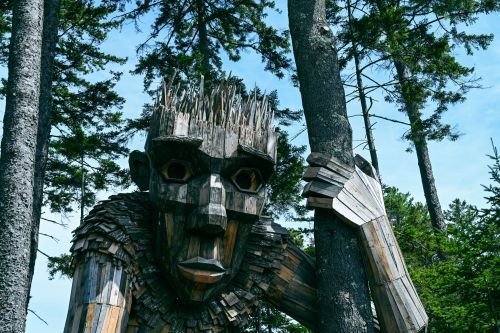 A large wooden troll sculpture stands among tall trees, with intricate details and a whimsical expression.