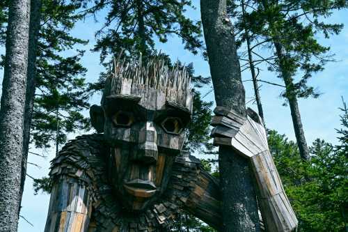 A large wooden troll sculpture stands among tall trees, with intricate details and a whimsical expression.