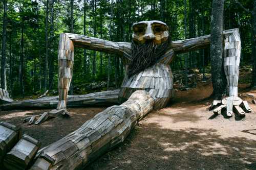 A large wooden sculpture of a giant figure with a beard, sitting among trees in a forested area.