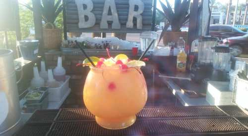 A large, colorful cocktail garnished with cherries and lime slices sits on a bar counter with a "BAR" sign in the background.