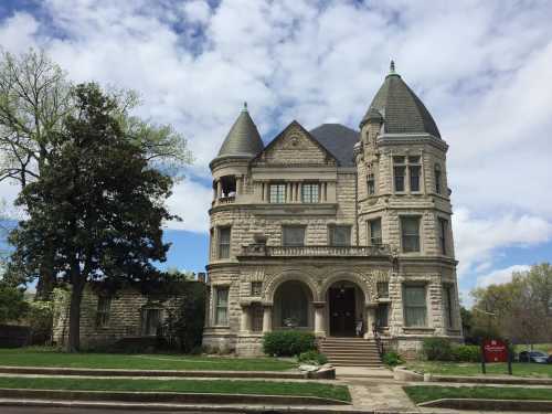 A large, ornate stone mansion with turrets and a landscaped front yard under a partly cloudy sky.