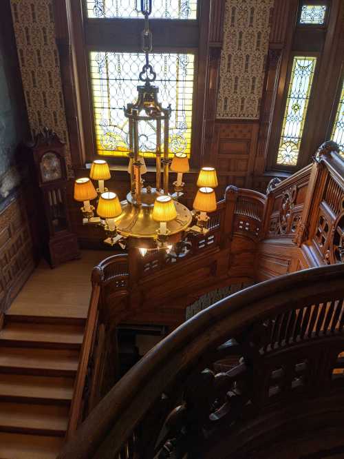 A grand wooden staircase with an ornate chandelier and stained glass windows, showcasing intricate architectural details.