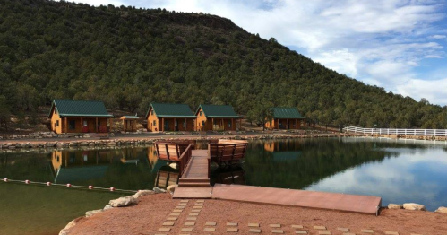 A serene landscape featuring wooden cabins by a calm lake, surrounded by green hills and a clear blue sky.