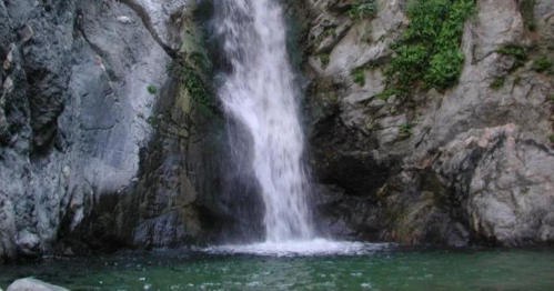 A serene waterfall cascading over rocky cliffs into a clear green pool below, surrounded by lush greenery.