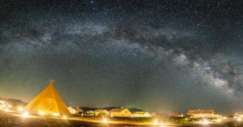 A starry night sky with the Milky Way above a lit campsite featuring a teepee and nearby houses.
