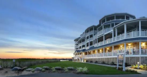 A coastal hotel at sunset, featuring multiple balconies and lush green landscaping in the foreground.