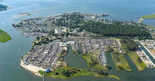 Aerial view of a coastal area with a marina, parking lots, and green spaces surrounded by water.