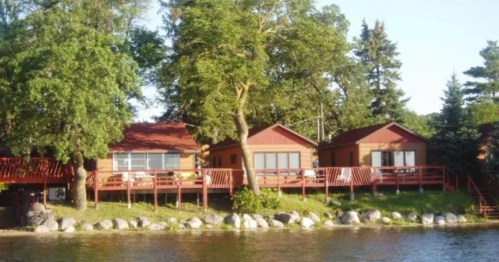 A serene lakeside view featuring wooden cabins with red accents, surrounded by trees and a rocky shoreline.