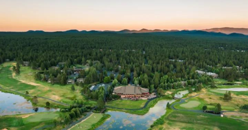 Aerial view of a golf course surrounded by lush trees and mountains at sunset, with a serene lake nearby.