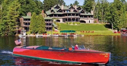 A vintage boat glides across a lake in front of a large, rustic mansion surrounded by trees.
