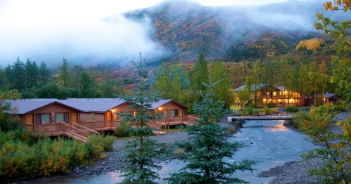 A serene landscape featuring cozy cabins by a river, surrounded by trees and misty mountains at dusk.