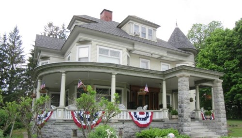 A large, elegant house with a wraparound porch, decorated with American flags and bunting, surrounded by greenery.