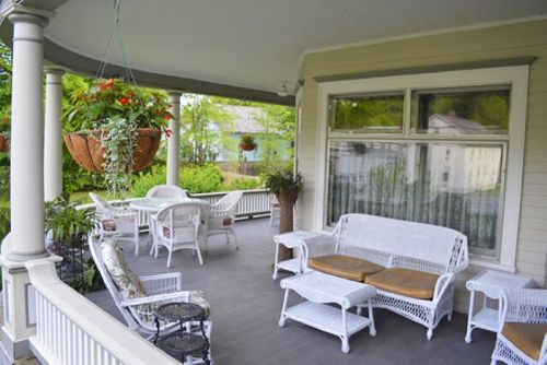 A cozy porch with white wicker furniture, potted plants, and hanging flower baskets, surrounded by greenery.
