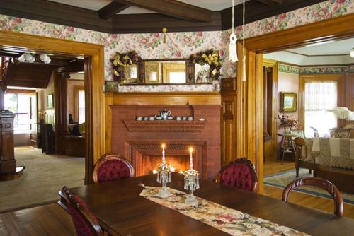 A cozy dining room with a wooden table, candles, and floral wallpaper, leading to a warm, inviting living space.