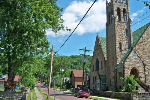 A scenic street view featuring a stone church, trees, and power lines under a blue sky with fluffy clouds.