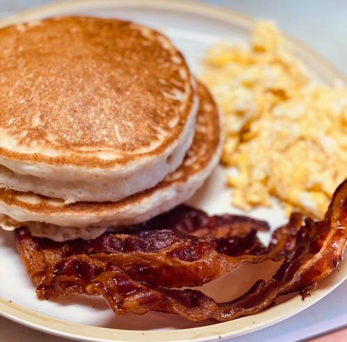 A plate with two fluffy pancakes, crispy bacon, and scrambled eggs.