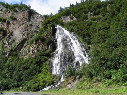 A cascading waterfall flows down a rocky cliff, surrounded by lush green vegetation and trees.