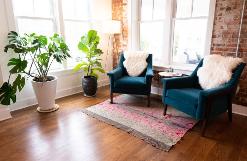 Cozy living space with two teal chairs, a plant, and a colorful rug against a brick wall and large windows.