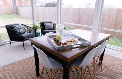 A cozy sunroom with a wooden table, a tray of snacks, and two black chairs beside a grassy outdoor view.