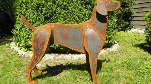 A rusted metal sculpture of a dog standing in a grassy area, surrounded by green bushes.