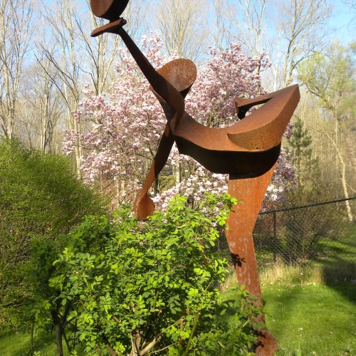A rusted metal sculpture of a bird stands among green foliage and blooming trees in a serene outdoor setting.
