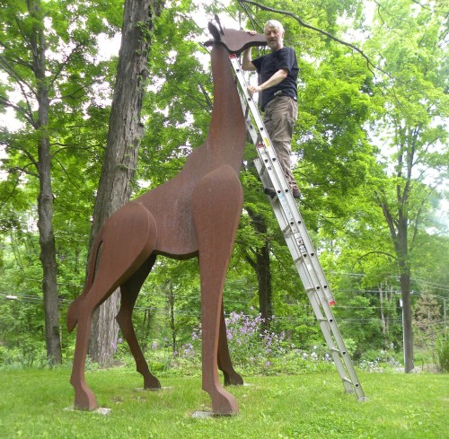 A person climbs a ladder next to a large metal giraffe sculpture in a green, wooded area.