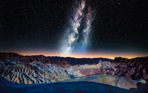 A stunning night sky filled with stars and the Milky Way over a rugged, colorful mountain landscape.