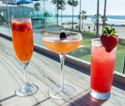 Three colorful cocktails on a marble table, with a beach view and palm trees in the background.