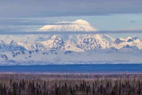 Snow-capped mountain peaks rise above a forested landscape under a cloudy sky.