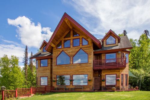 A large wooden house with multiple windows and a triangular roof, surrounded by greenery and blue skies.