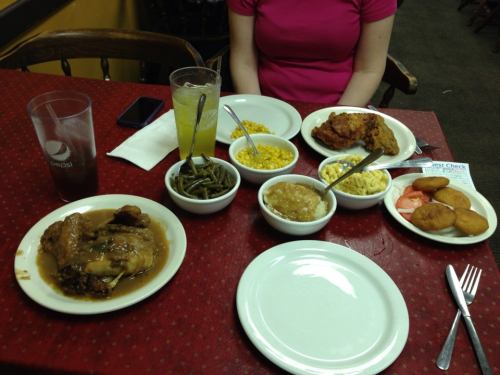 A table set with plates of fried chicken, sides of green beans, corn, macaroni, and desserts, with a drink nearby.