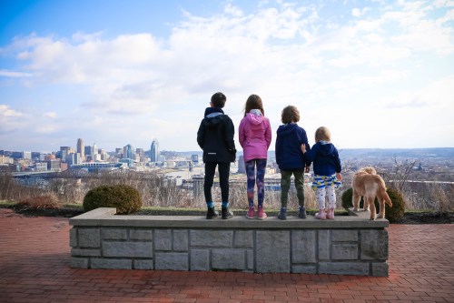 Four children and a dog stand on a stone wall, gazing at a city skyline under a cloudy sky.