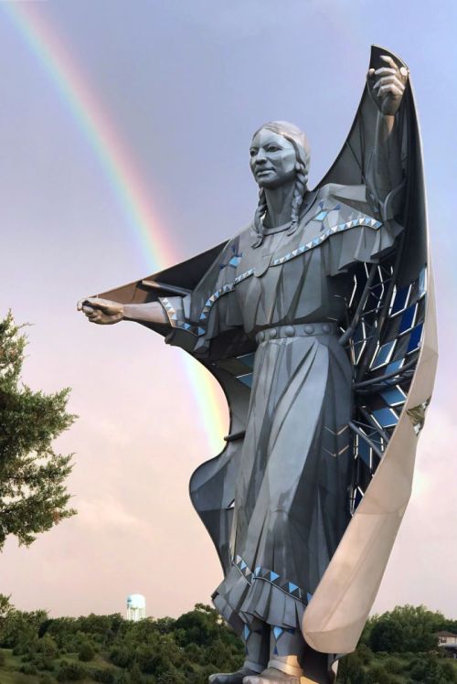 A large metallic statue of a woman with a rainbow in the background, showcasing intricate details and a flowing garment.