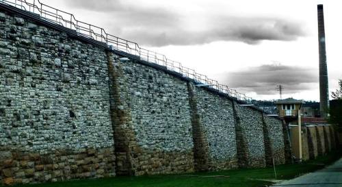 A tall stone wall with a metal railing, under a cloudy sky, next to a building and a tall chimney in the background.