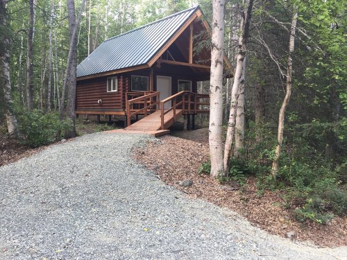 A small wooden cabin with a sloped roof, surrounded by trees and a gravel path leading to the entrance.