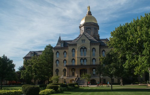Historic building with a golden dome, surrounded by green trees and a well-maintained lawn under a blue sky.