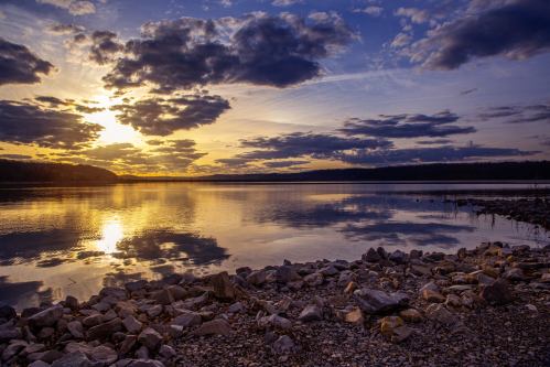 A serene sunset over a calm lake, with clouds reflecting on the water and rocky shore in the foreground.