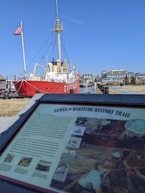 A red lighthouse ship docked near a sign about the Lewes Maritime History Trail under a clear blue sky.
